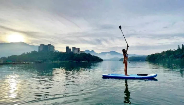 有人站在湖面的立槳板上舉槳,背景是山與湖岸建築