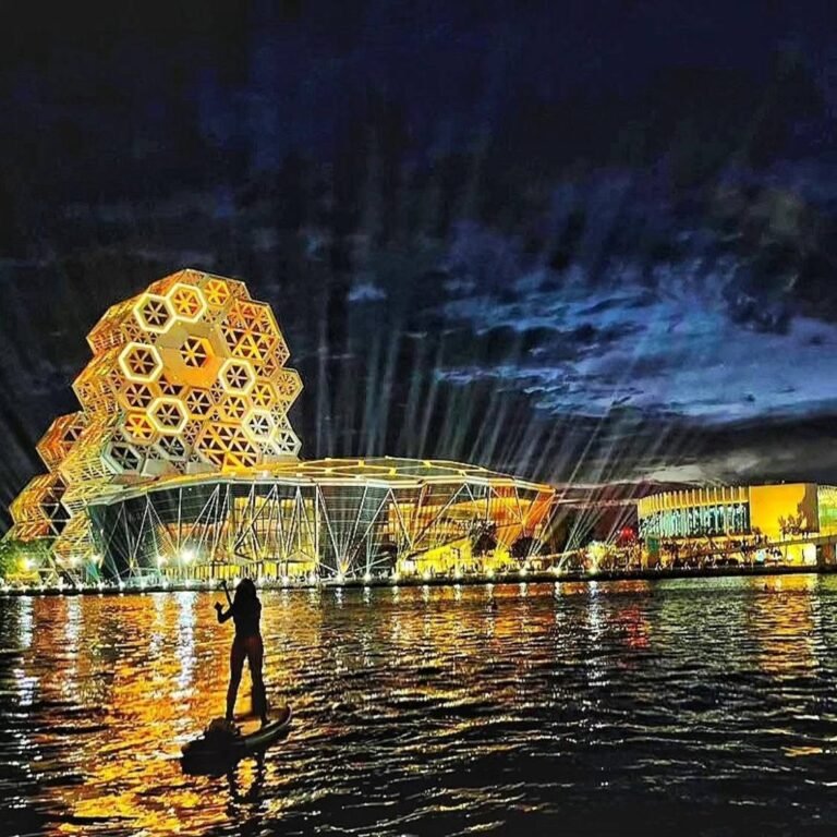 夜晚有人站在立槳板上，背景是燈光亮起的金色蜂巢造型建築