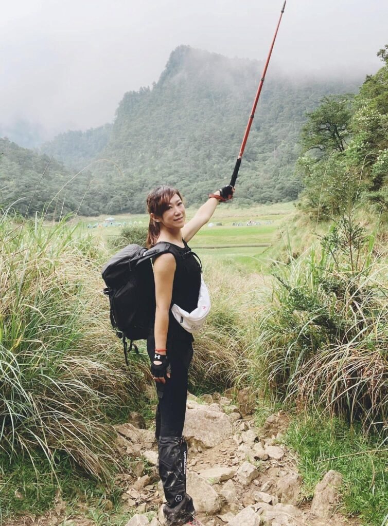 女生背著背包、舉著登山杖,站在雲霧繚繞的山區草徑上