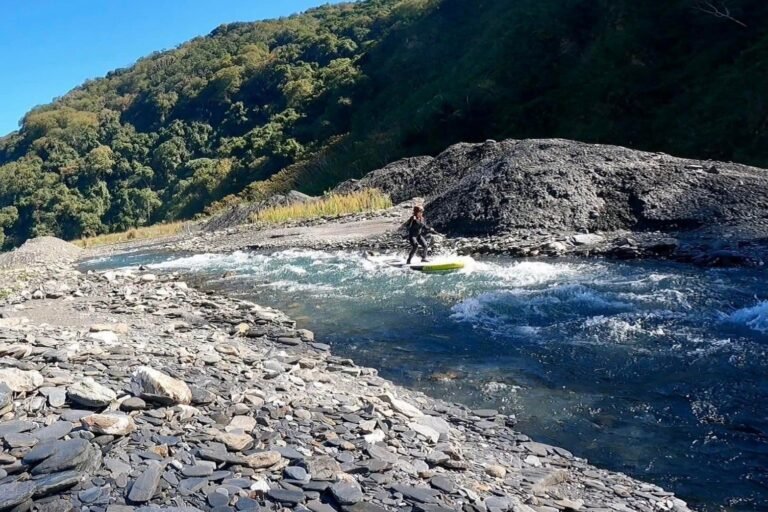 有人站在立槳板上通過溪谷急流,兩岸是碎石灘與青山