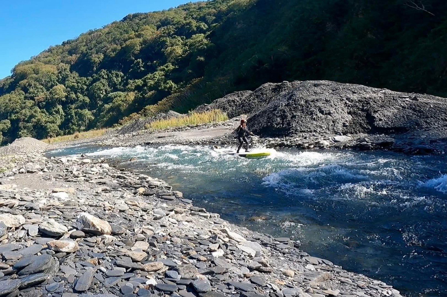 有人站在立槳板上通過溪谷急流,兩岸是碎石灘與青山