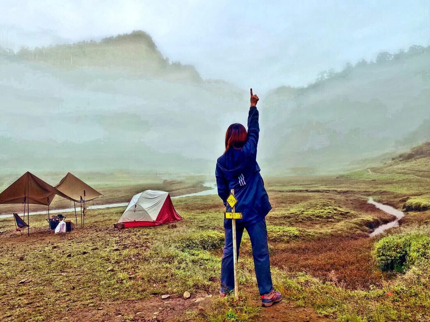 霧氣瀰漫的山谷草原上搭著帳篷,一位登山者舉手指向天空